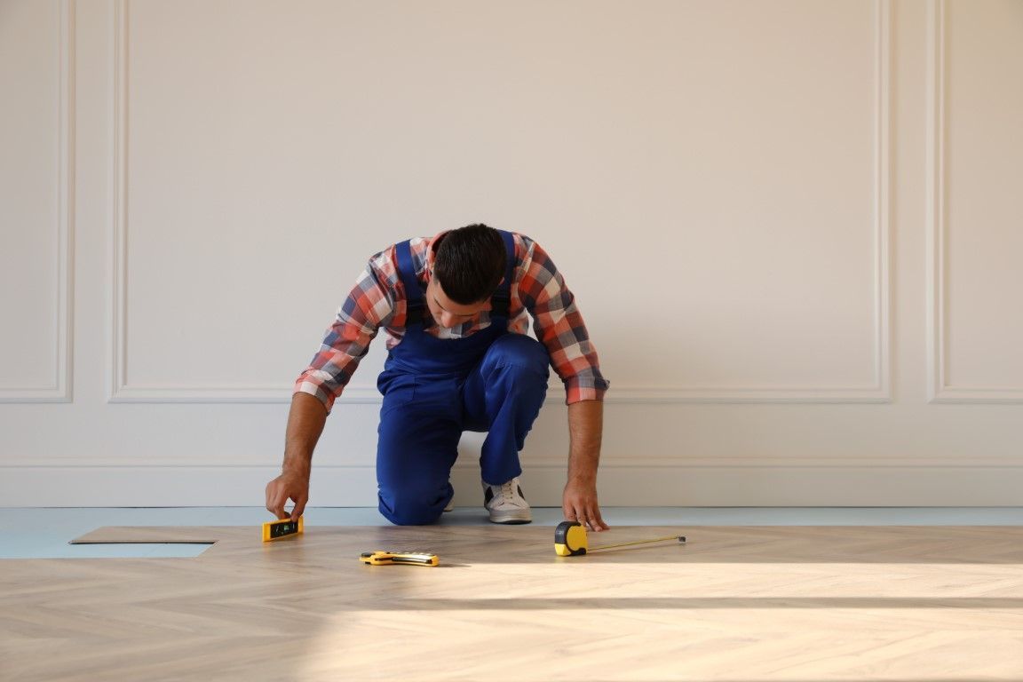 Person kneeling on a wooden floor, measuring and marking planks with yellow tools in a bright room