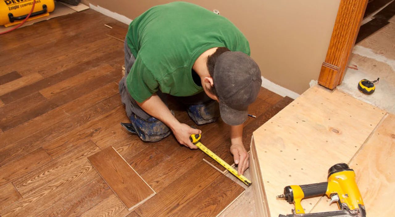 Man kneeling on a wood floor, measuring and marking a plank with a tape measure during installation.