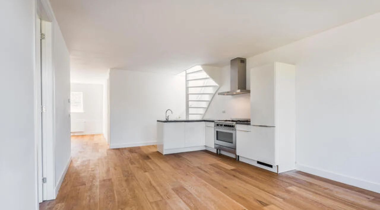 Bright empty kitchen and living area with hardwood floors, white walls, and stainless steel appliances