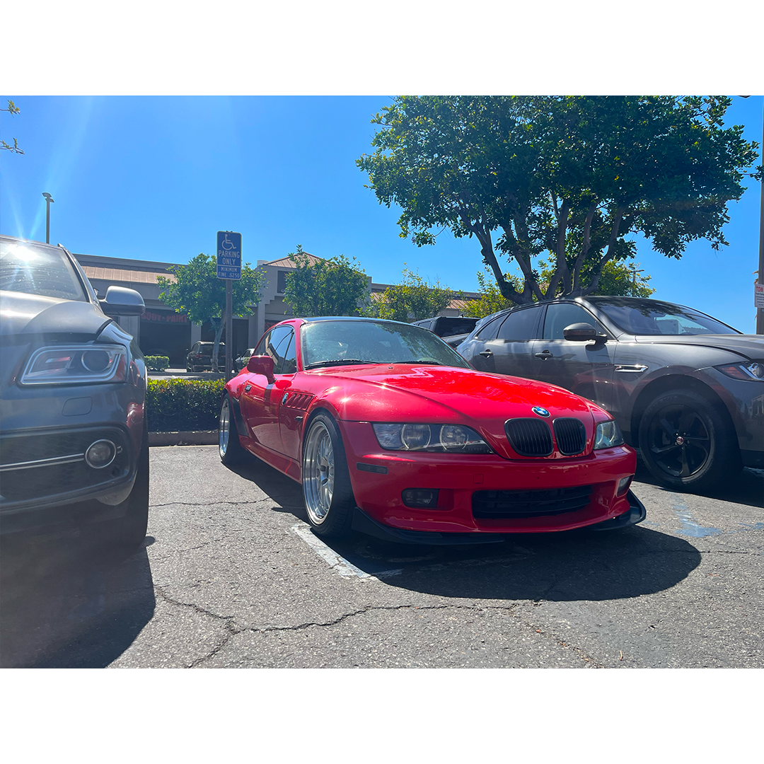 Red BMW sports car parked between two gray cars on a sunny street.