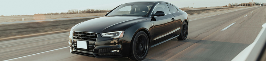 Black sedan driving on a highway, viewed from the front-left side.