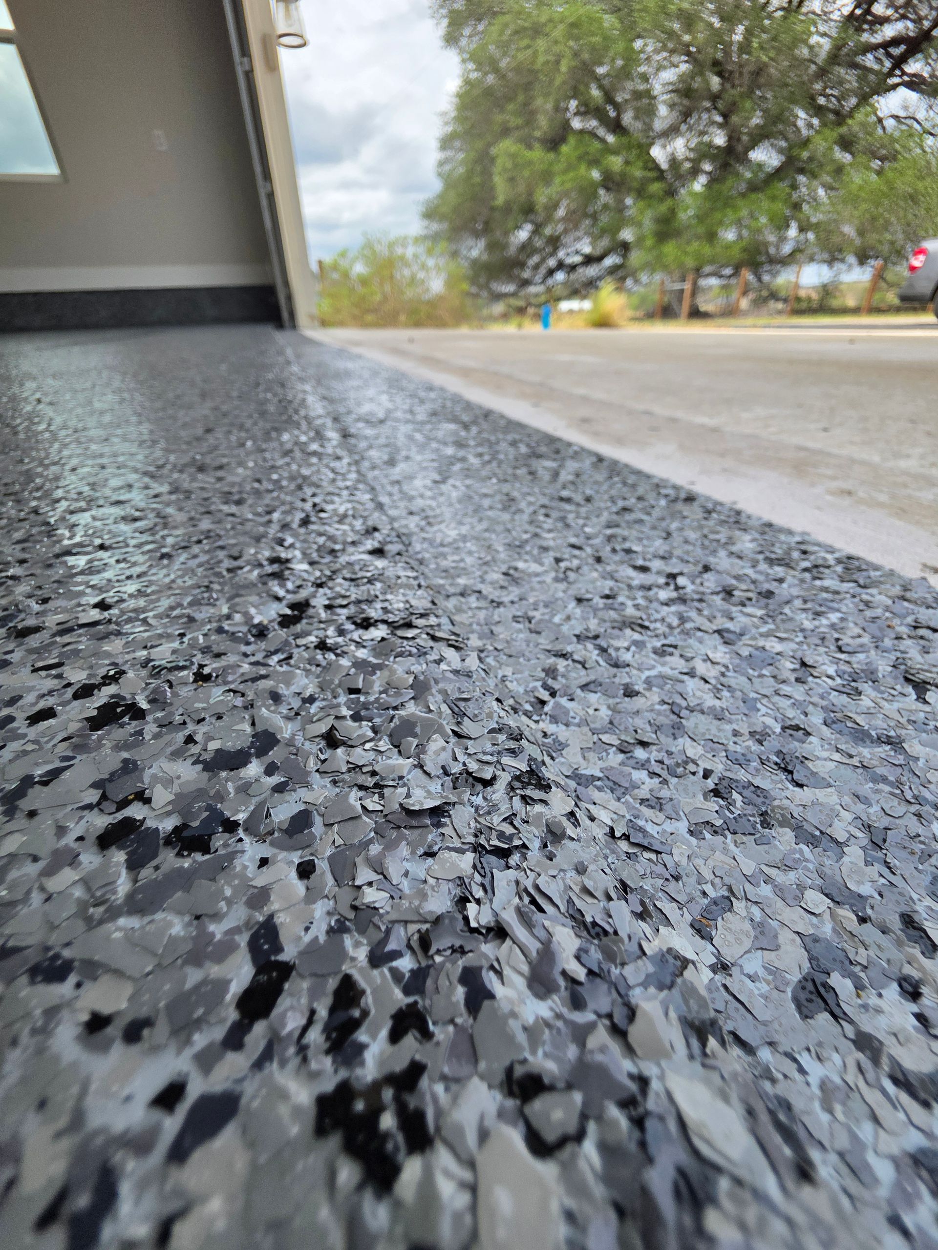 Low-angle view of a speckled black garage floor leading to a tree-lined driveway outdoors