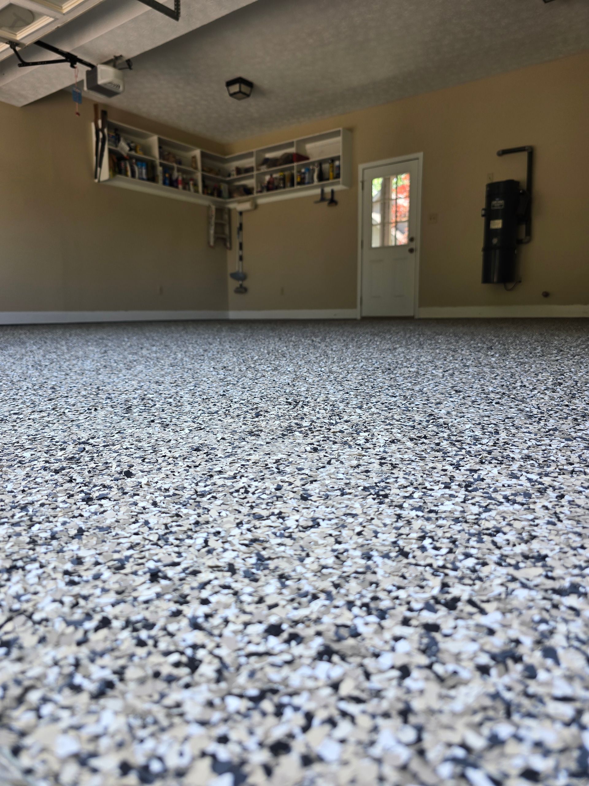 Empty garage interior with speckled epoxy floor, beige walls, built-in shelves, and a side door.