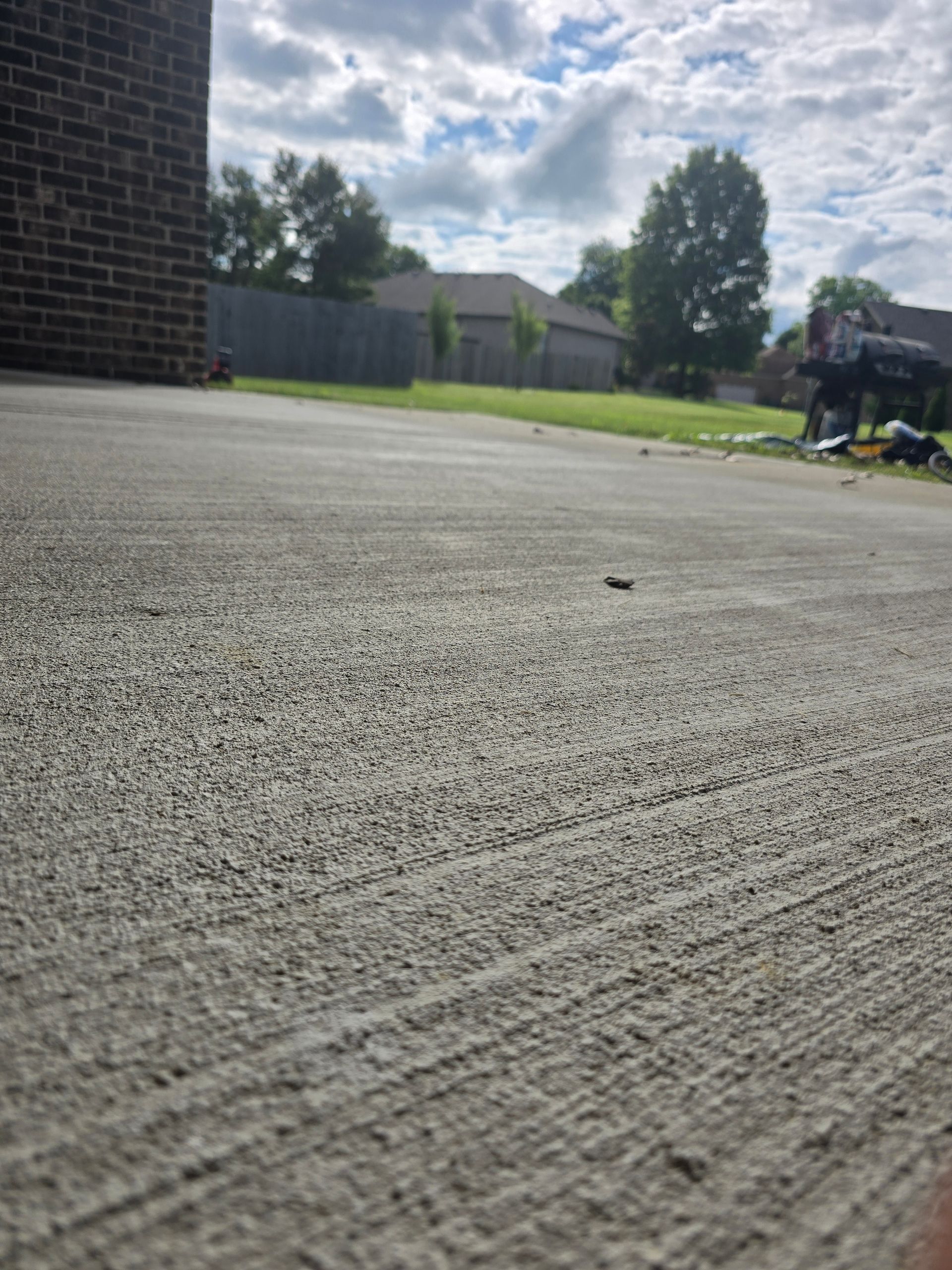 Low-angle view of a concrete driveway with a fence, trees, and a house in the background.