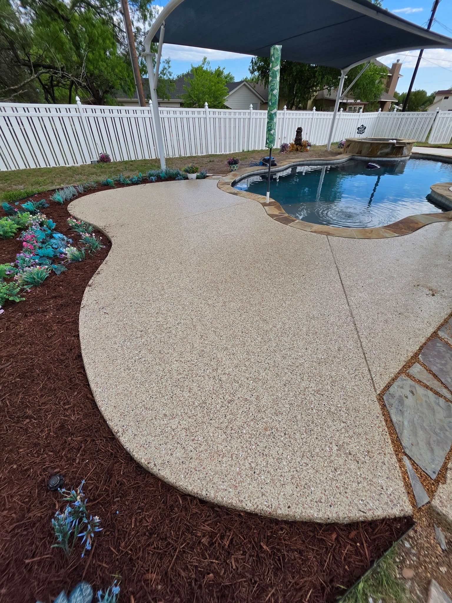 Backyard poolside patio with pebble concrete, mulch beds, white fence, and shade sail