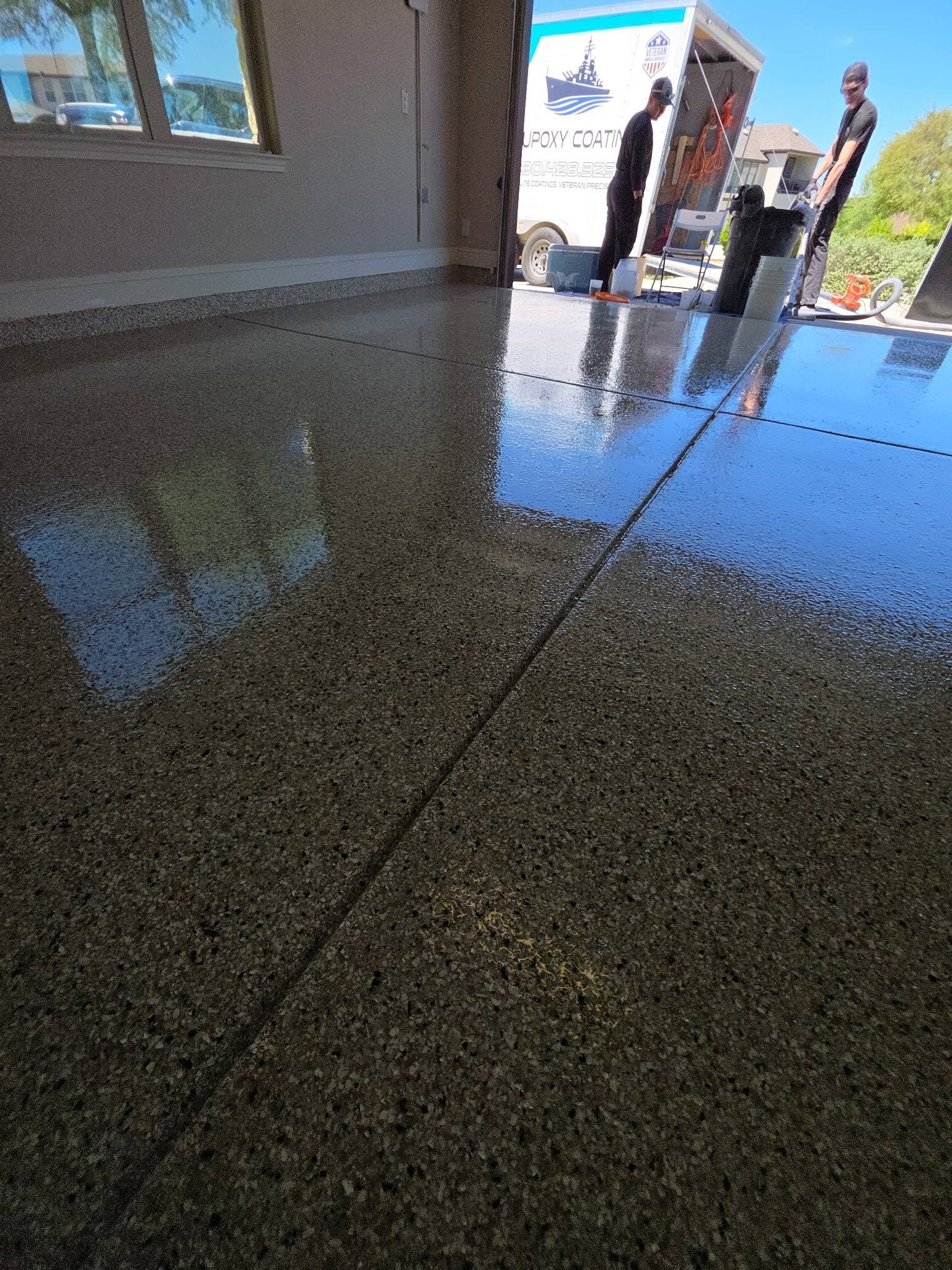 Sunlit wet concrete walkway outside a building, with two people near the entrance and reflections on the floor