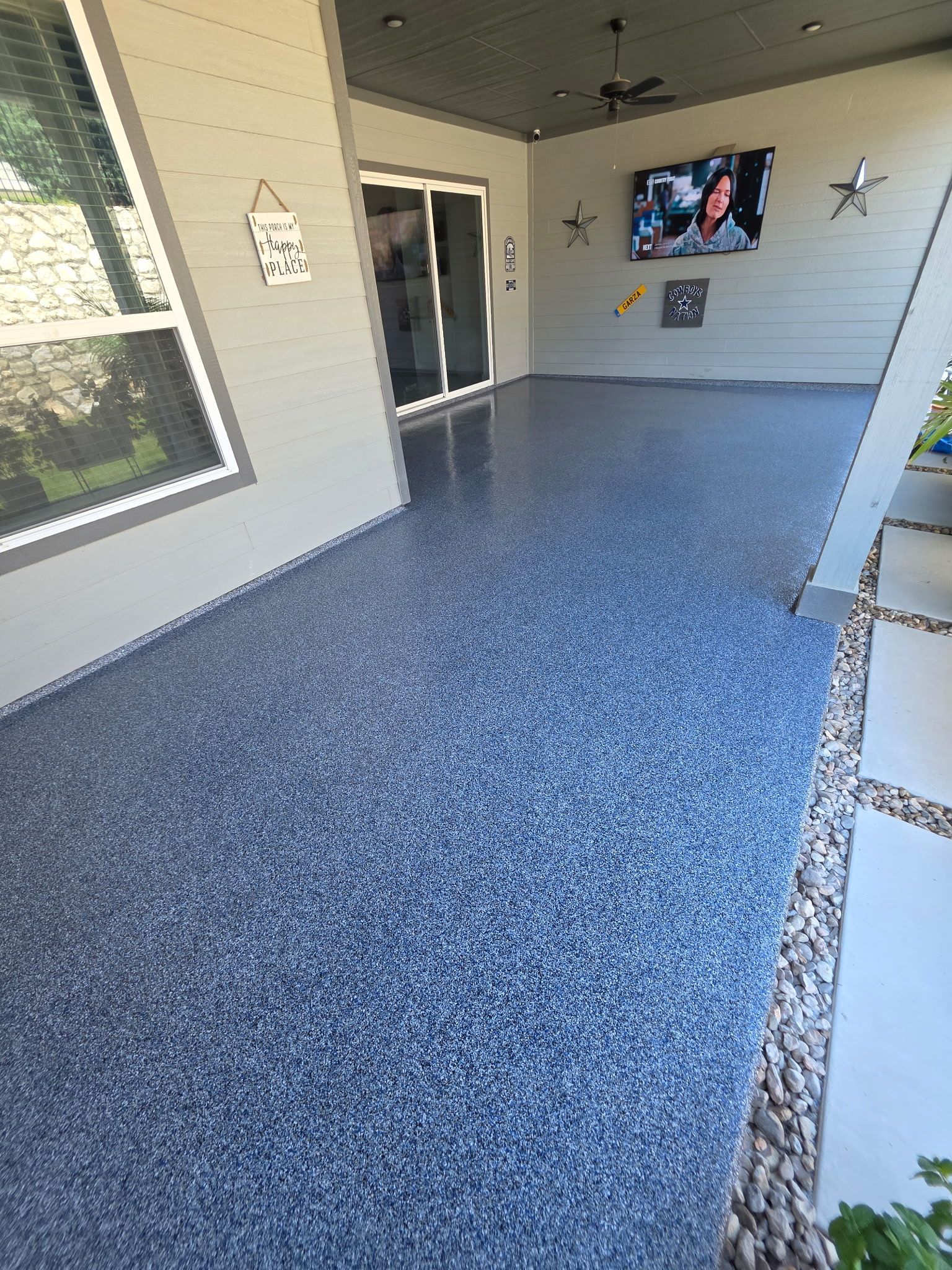 Covered patio with speckled blue floor, ceiling fan, wall-mounted TV, and sliding glass door.