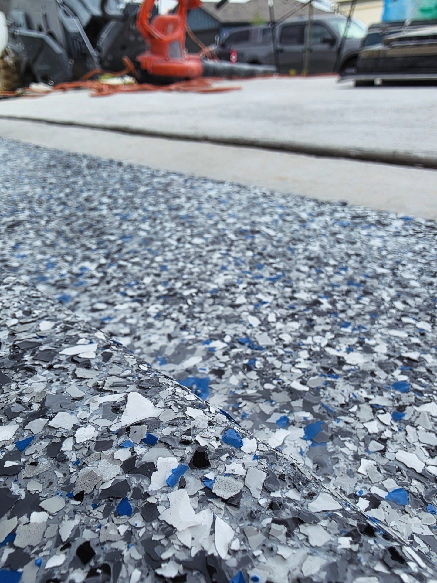 Close-up of speckled gray epoxy floor with blue flecks in a garage or workshop, blurred tools in background