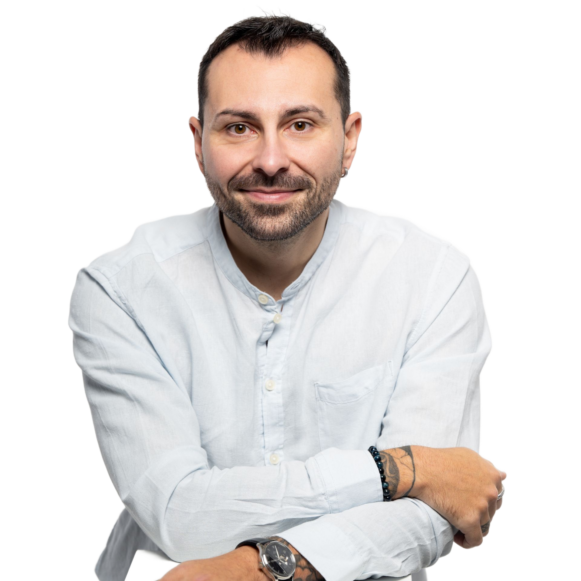 Homme aux cheveux courts et foncés, souriant, portant une chemise bleu clair boutonnée, une montre et des bracelets, appuyé sur quelque chose de blanc.