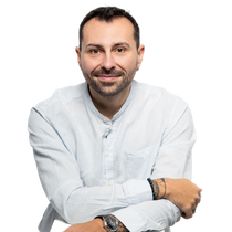 Homme aux cheveux courts et foncés, souriant, portant une chemise bleu clair boutonnée, une montre et des bracelets, appuyé sur quelque chose de blanc.
