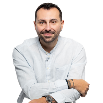 Homme aux cheveux courts et foncés, souriant, portant une chemise bleu clair boutonnée, une montre et des bracelets, appuyé sur quelque chose de blanc.