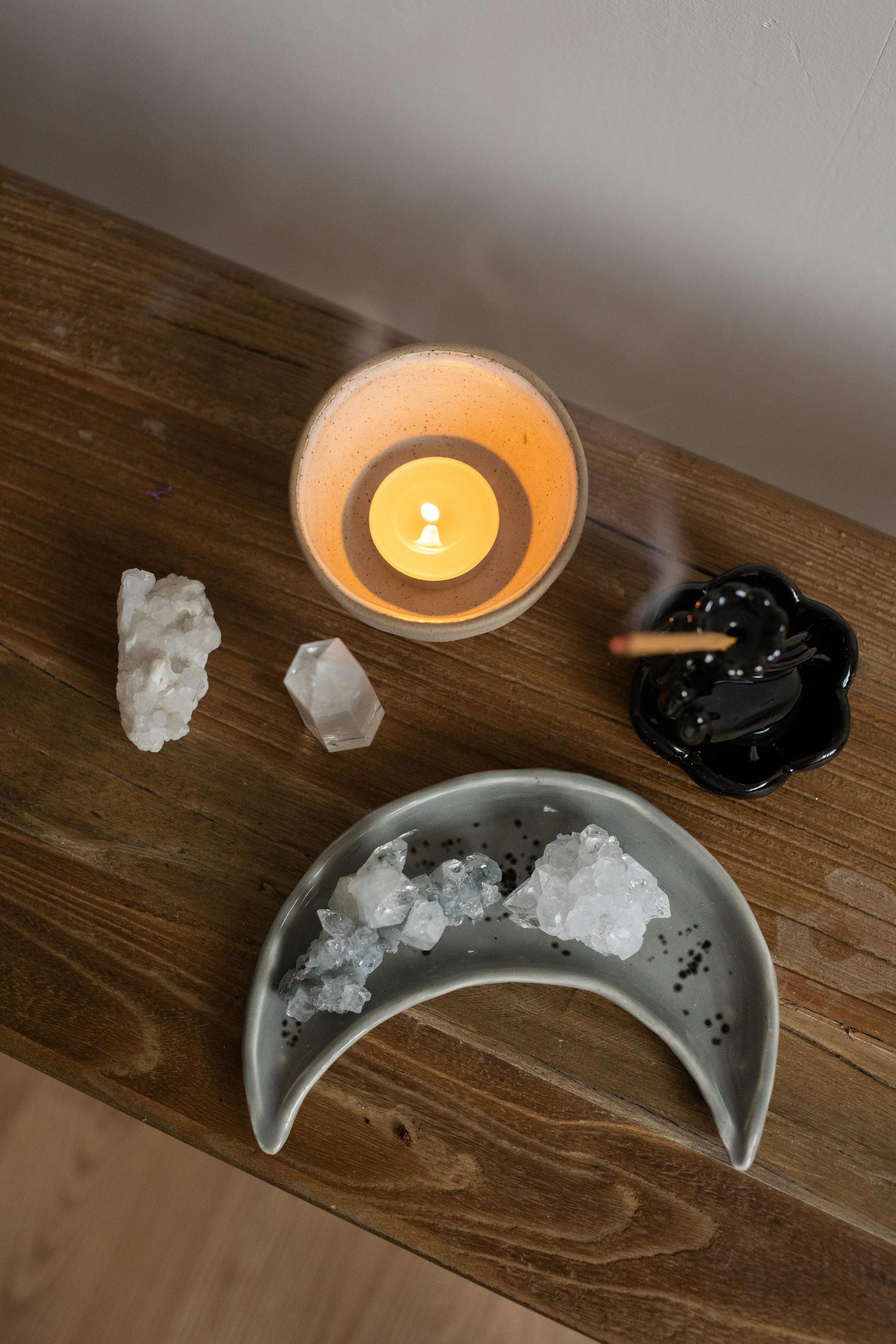 Étagère en bois avec une bougie allumée, des cristaux, un plateau en forme de lune avec d'autres cristaux et un porte-encens fumant.