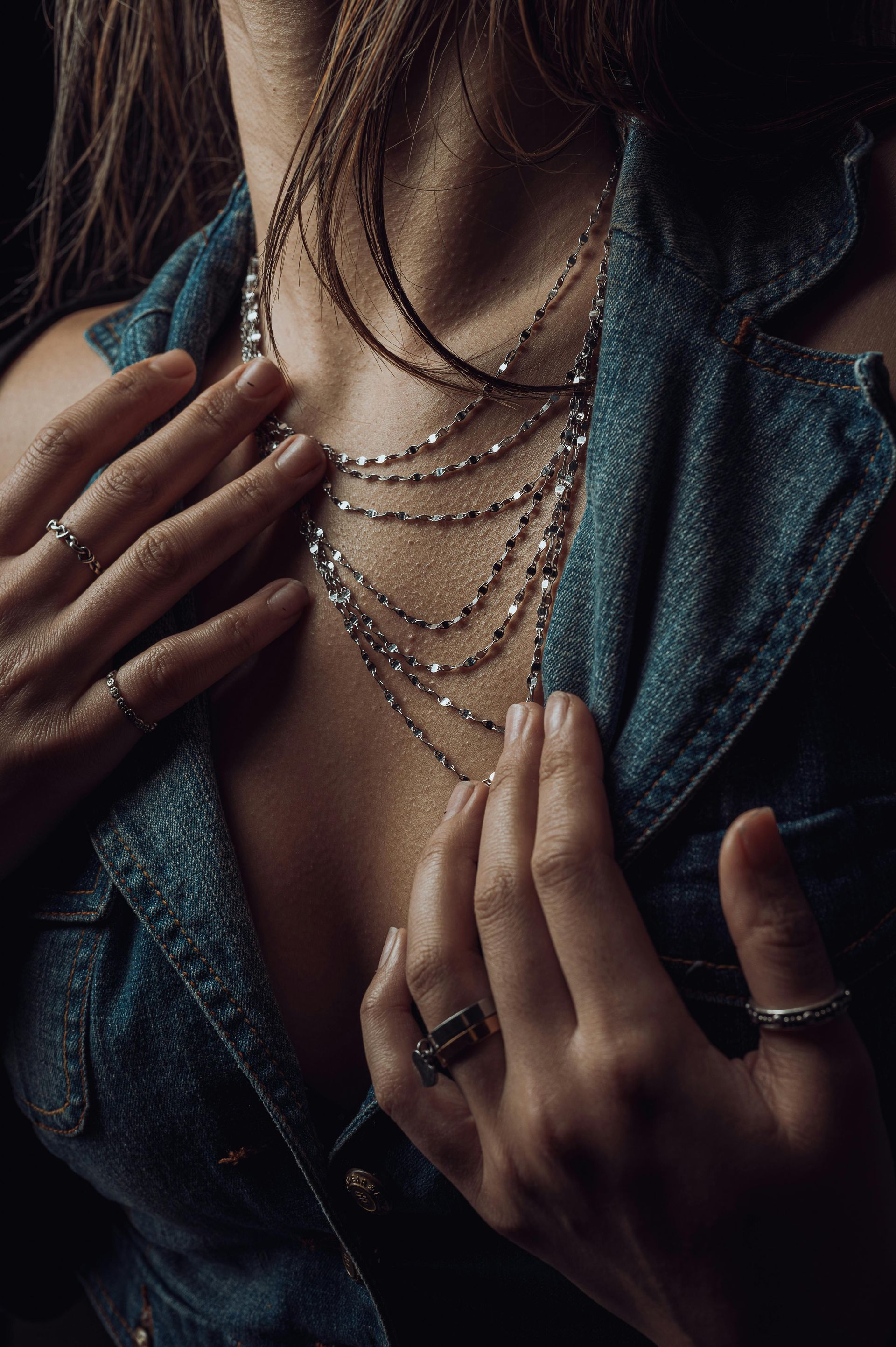 Femme portant plusieurs colliers superposés, une veste en jean, les mains près de la poitrine, des bagues aux doigts.