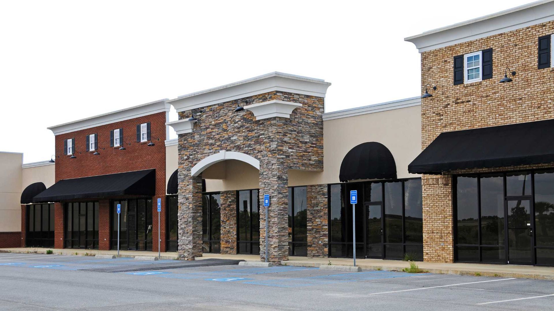 Empty strip mall storefronts with stone facades and black awnings in a parking lot
