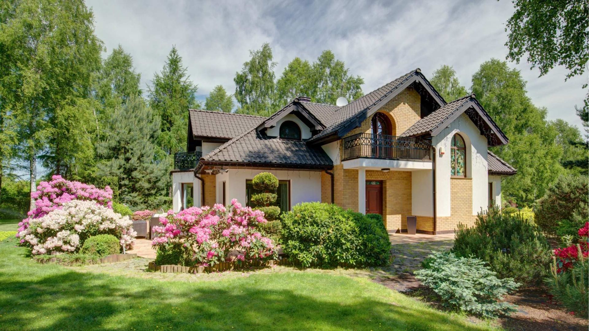A charming house with a garden of pink flowers, green shrubs, and lush lawn under a cloudy sky