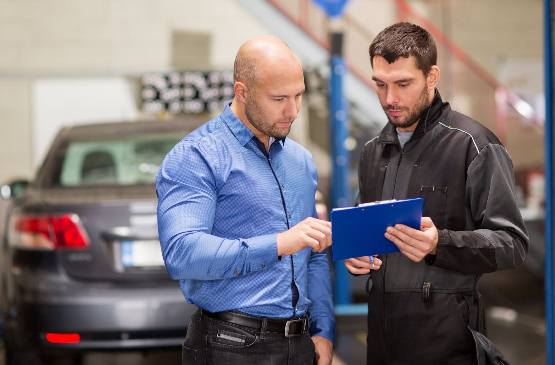 Man in blue shirt reviews paperwork with mechanic in a garage; car in the background. Man in blue shirt reviews paperwork with mechanic in a garage; car in the background.
