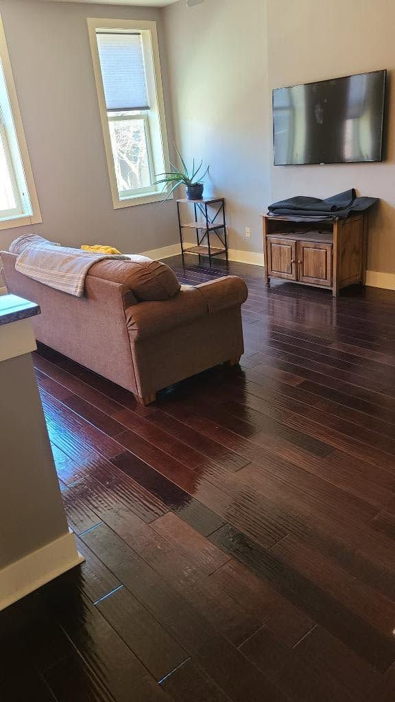 Bright living room with dark hardwood floors, a brown sofa, wall-mounted TV, and large windows.