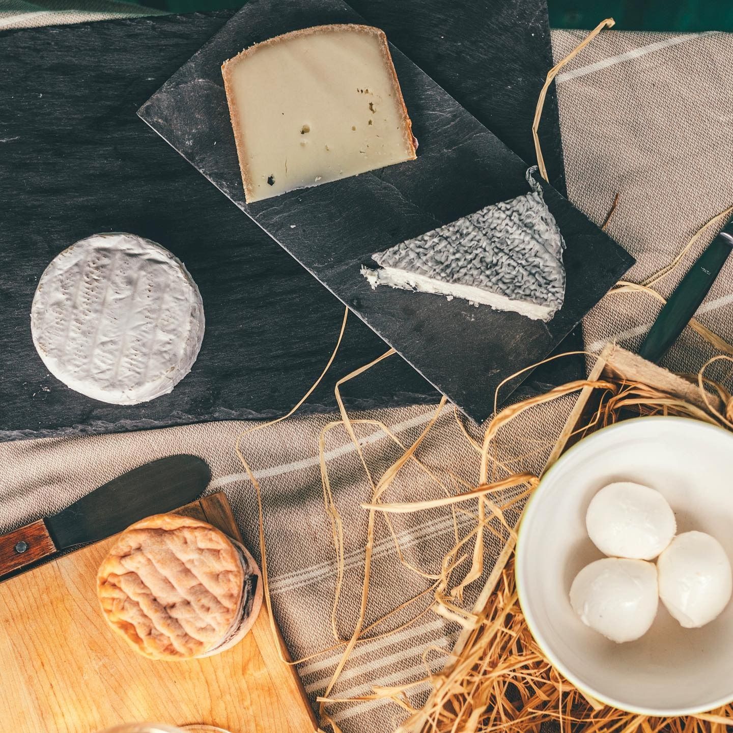 Fromages, crackers, pain et boules de fromage blanc disposés sur une table rustique sombre agrémentée de paille et de tissu.