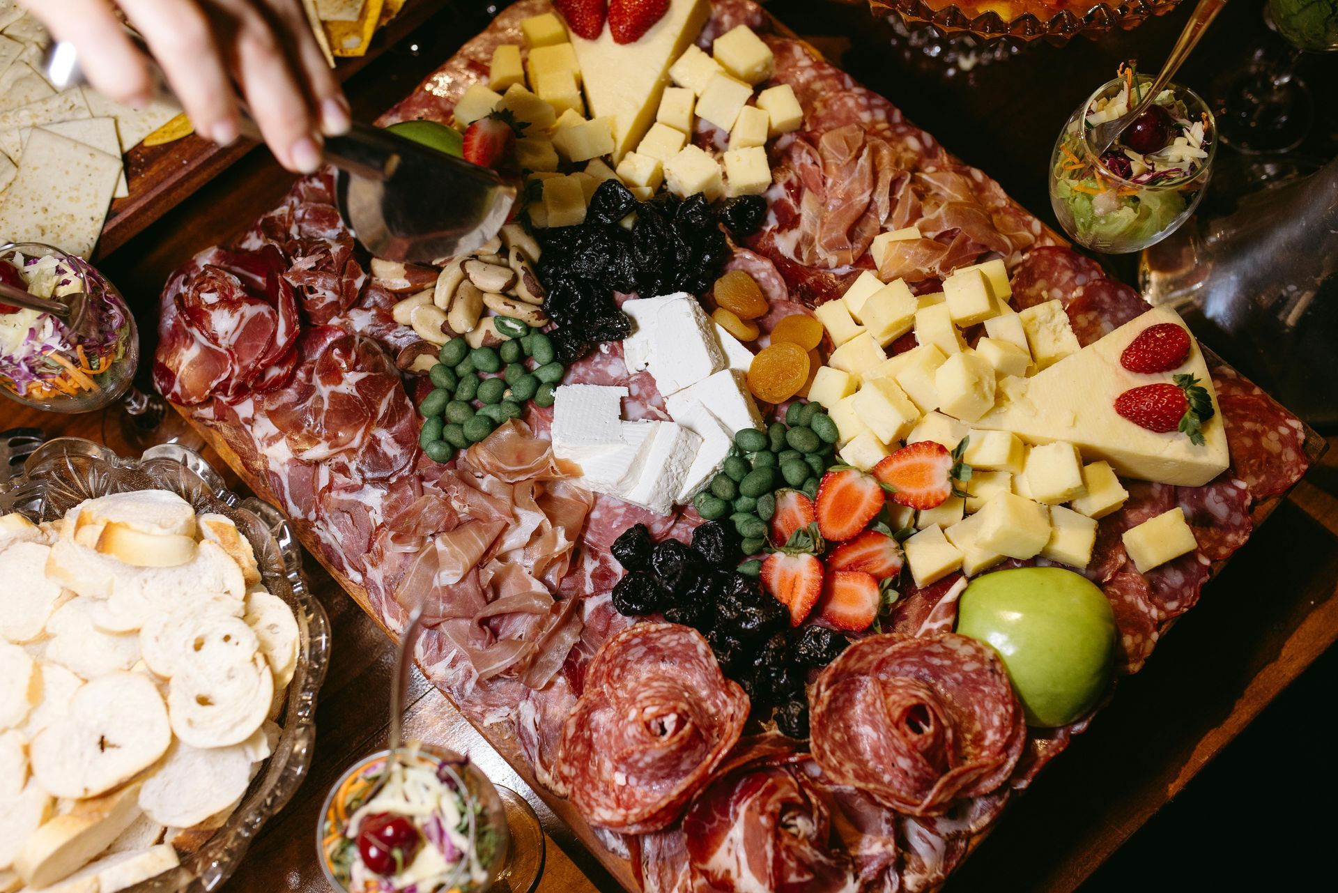 Planche de charcuterie avec fromages, viandes, raisins, fraises et tranches de pomme, avec un service à la main