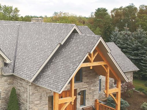 Stone house with gray shingle roof and timber porch under construction, surrounded by trees