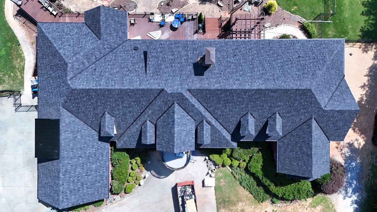 Aerial view of a large gray-roofed house with a central walkway and surrounding trees and patio.