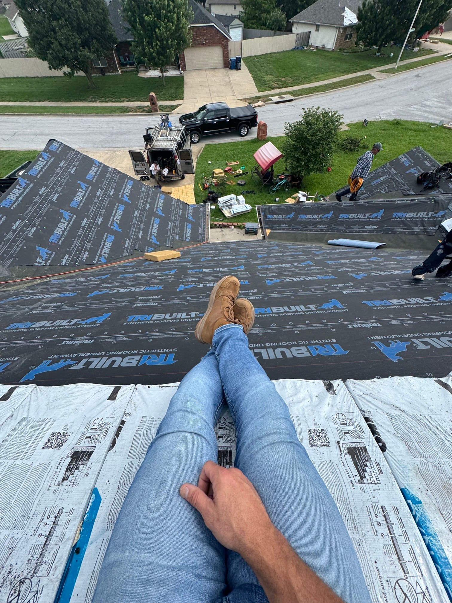 Person’s view from a steep roof, looking down at a house and driveway below with roofing underlayment laid out.