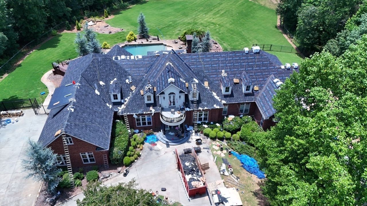 Aerial view of a large brick mansion with a dark roof, surrounded by trees and a lawn
