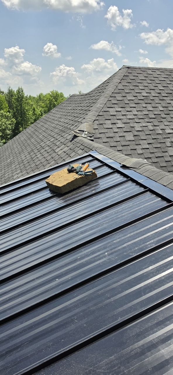 Roof with black metal panels and a small toolbox, backed by gray shingles and blue sky.