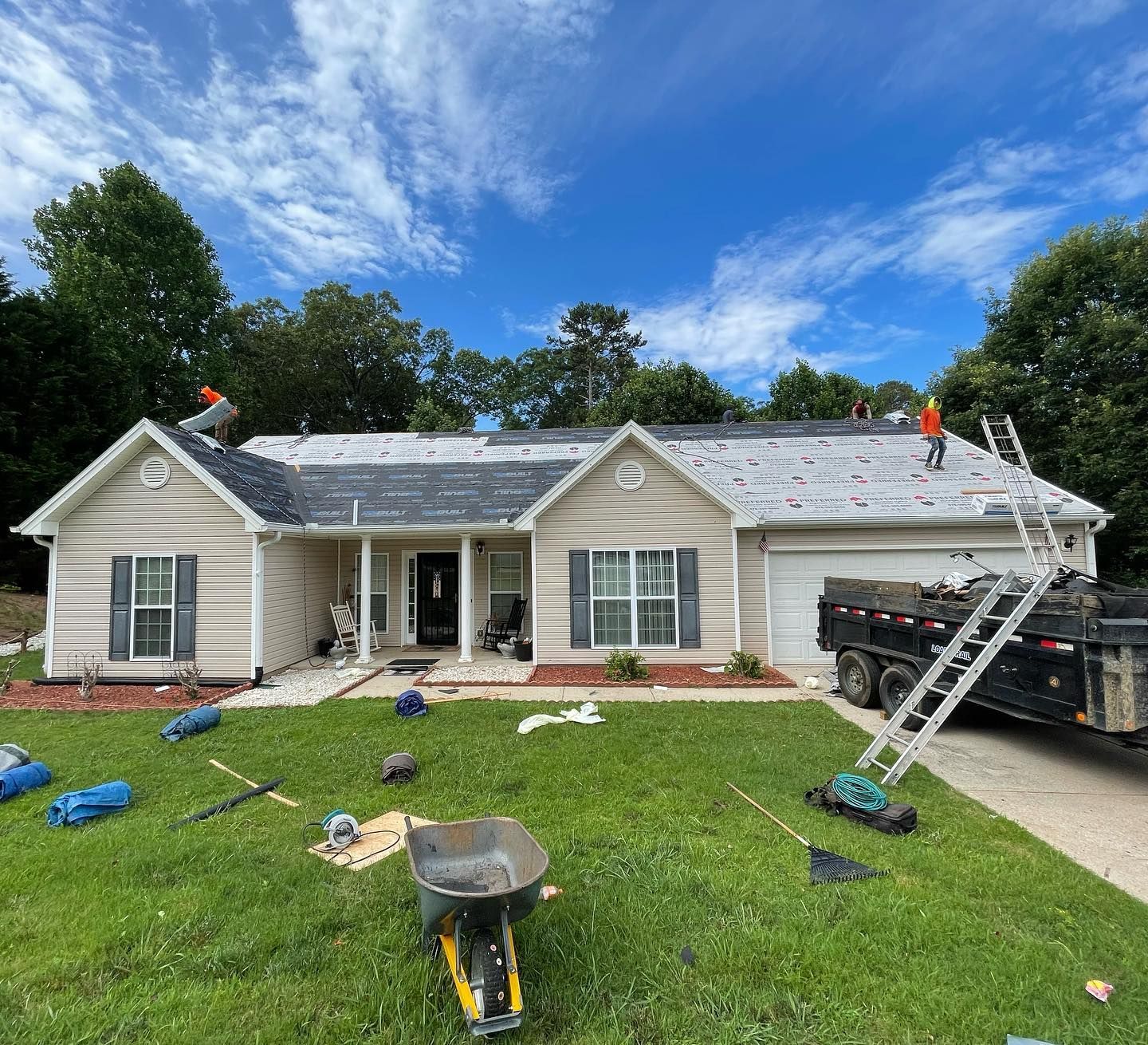 Roofing crew replacing shingles on a single-story house with ladders, tools, and scattered materials in the yard