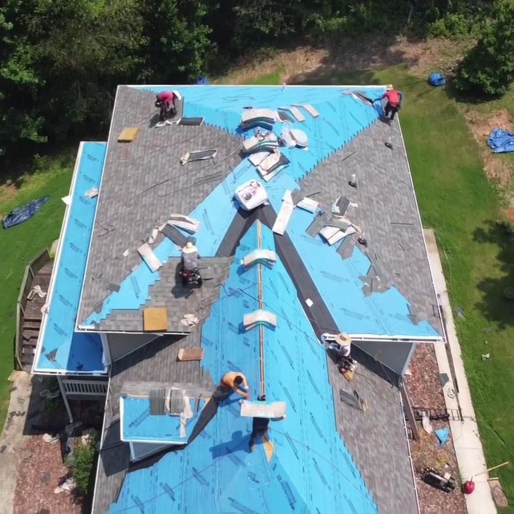 Workers repairing a roof with blue underlayment and gray shingles, seen from above