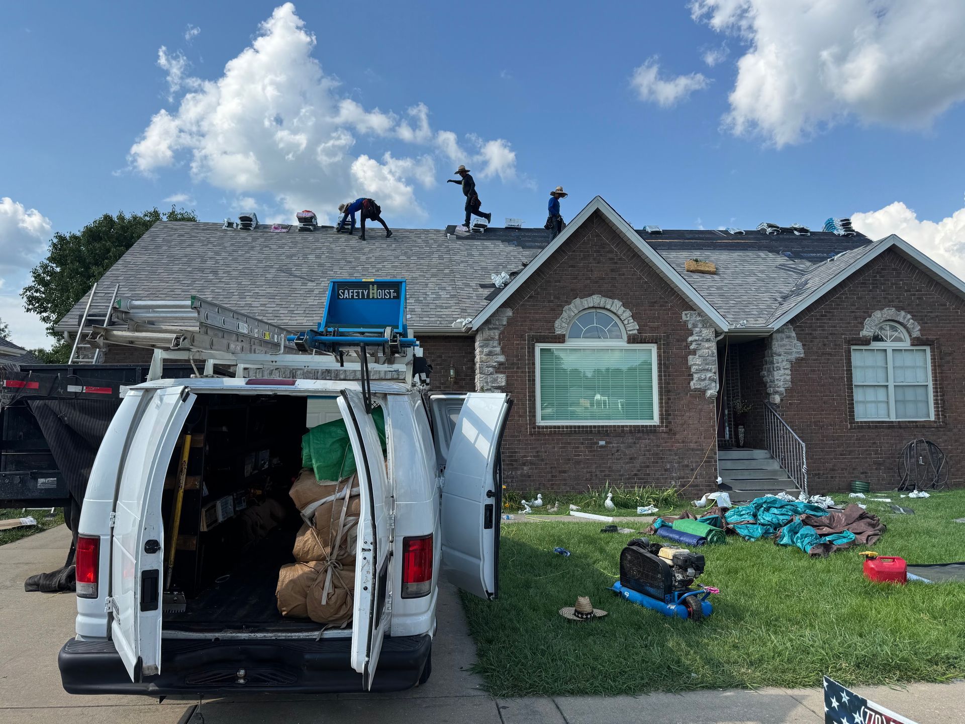 Workers on a roof beside a white van and storm debris in front of a brick house