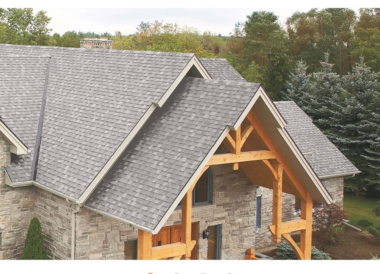 Stone house under construction with gray roof, wooden porch framing, and trees in the background