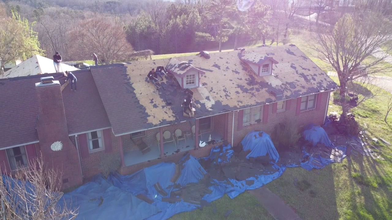 Aerial view of a brick house with a damaged roof in a wooded suburban yard.