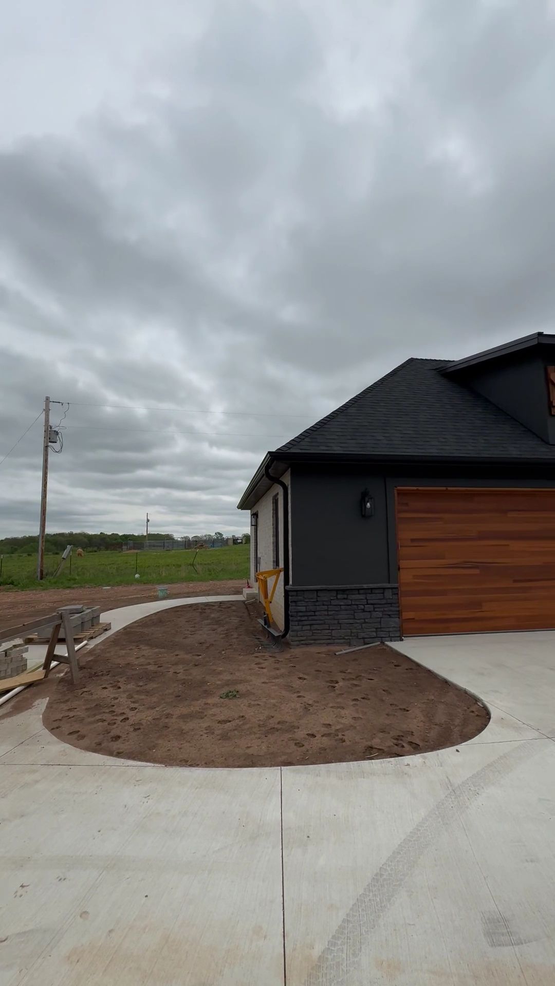 Modern house exterior with dark siding, wood garage door, and a newly landscaped front yard under cloudy skies