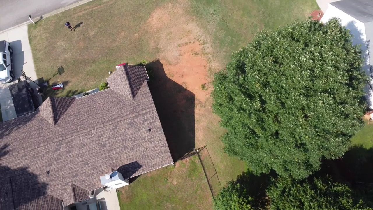 Aerial view of a house roof beside a grassy yard and large tree with a patch of bare dirt.