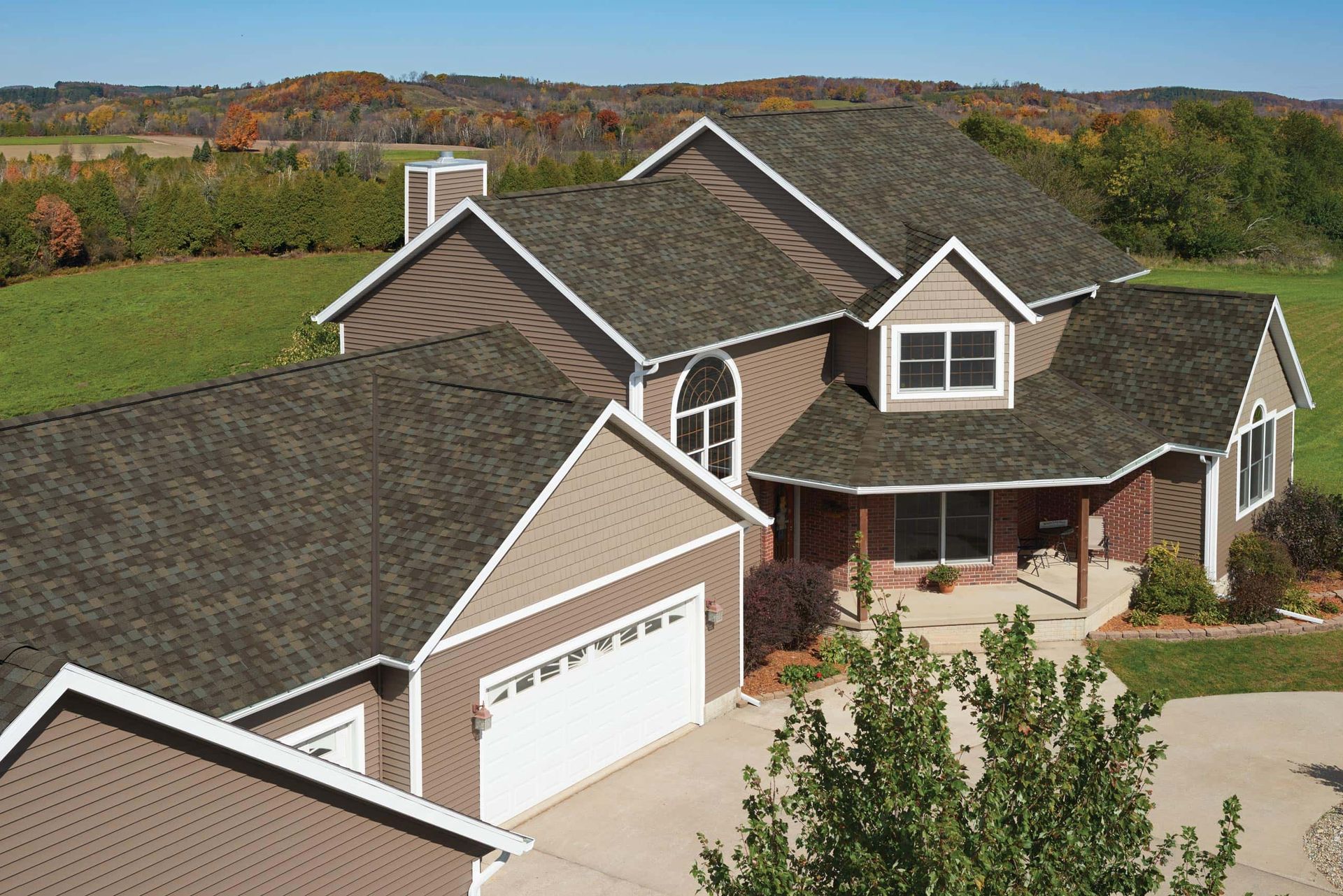 Aerial view of a large suburban house with a gray roof, garage, and front porch surrounded by trees.