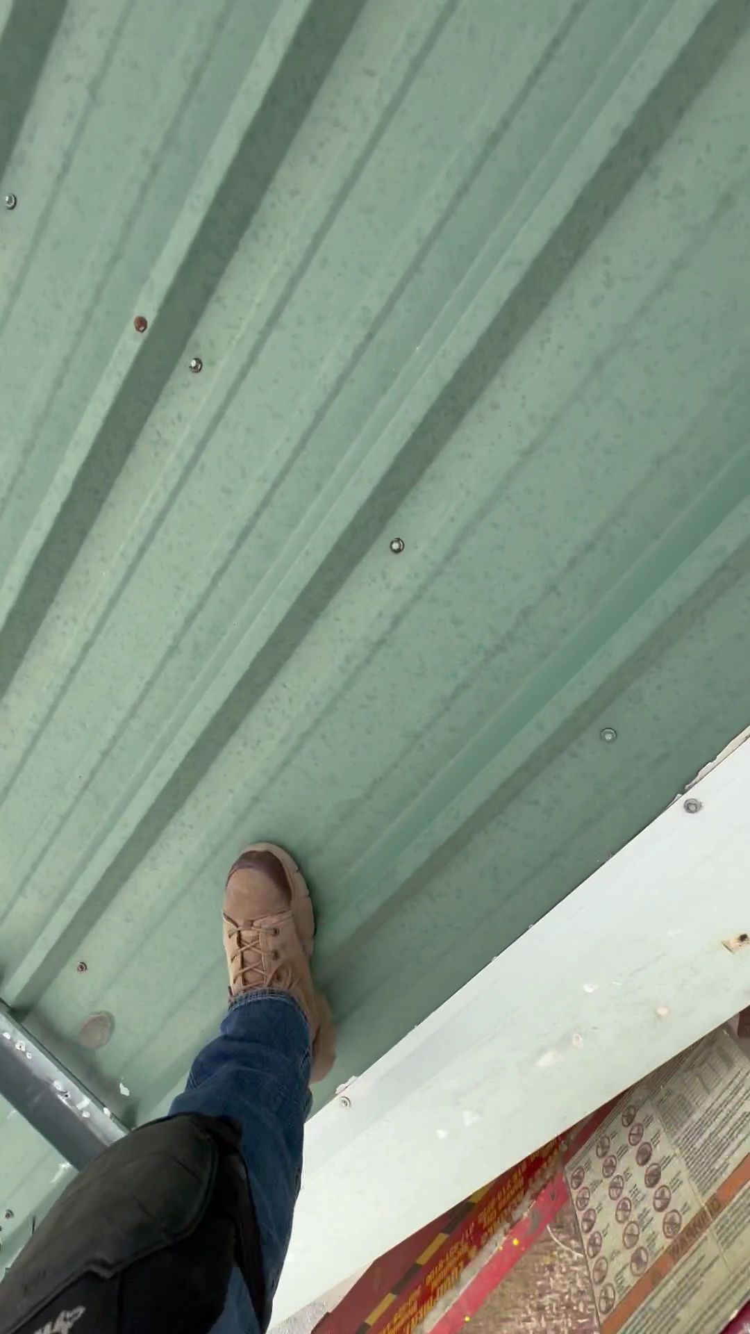A foot in a tan boot standing on a green corrugated metal roof edge.