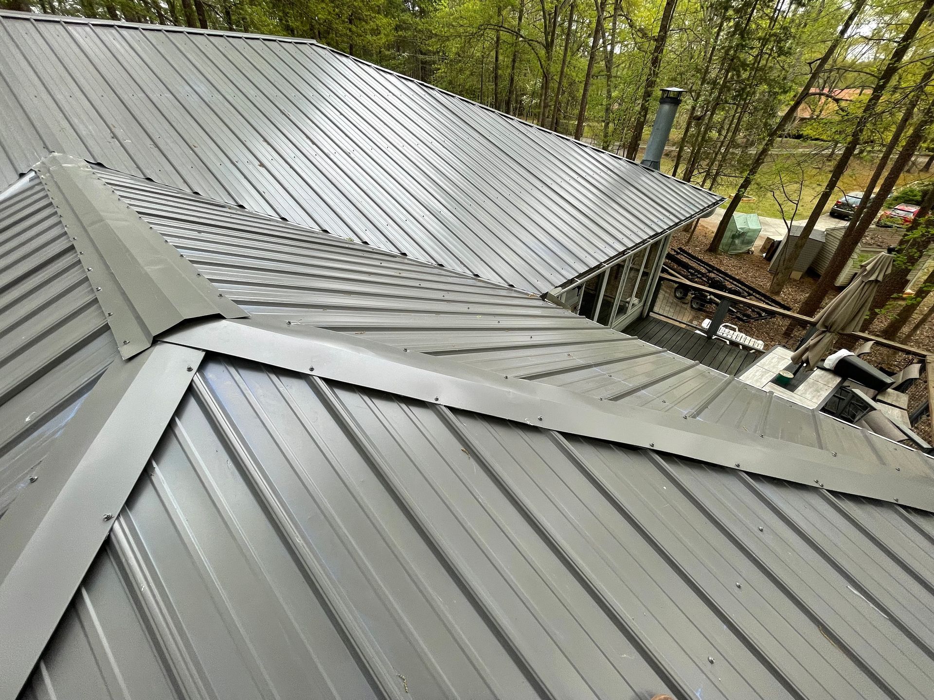 Gray metal roof with multiple slopes and chimneys, viewed from above with trees in the background
