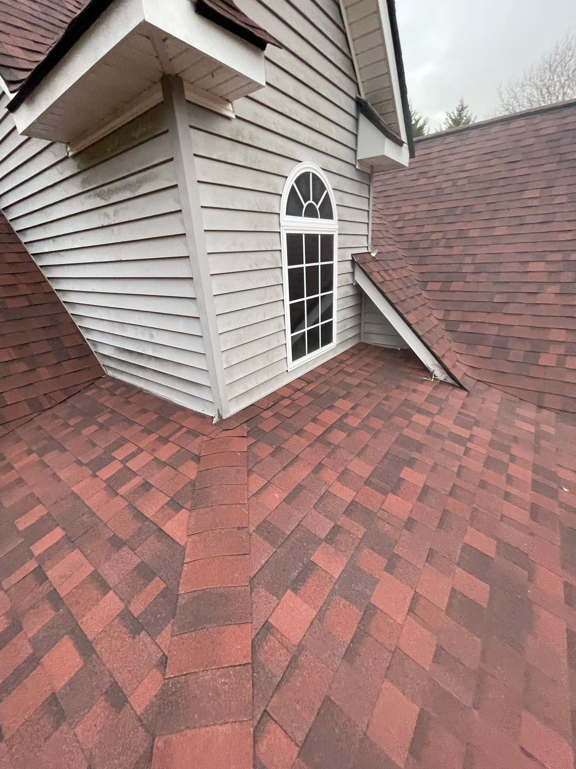 Red shingled roof with a dormer window and gray siding on a house exterior.