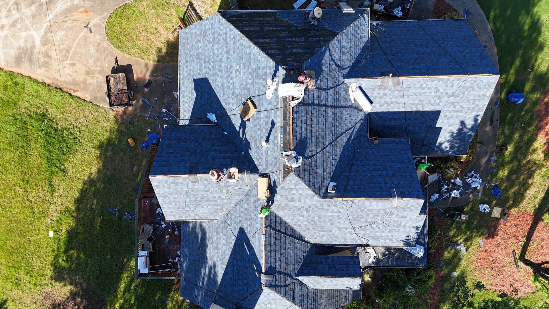 Aerial view of a gray-shingled house roof with chimneys, surrounded by lawn and trees.