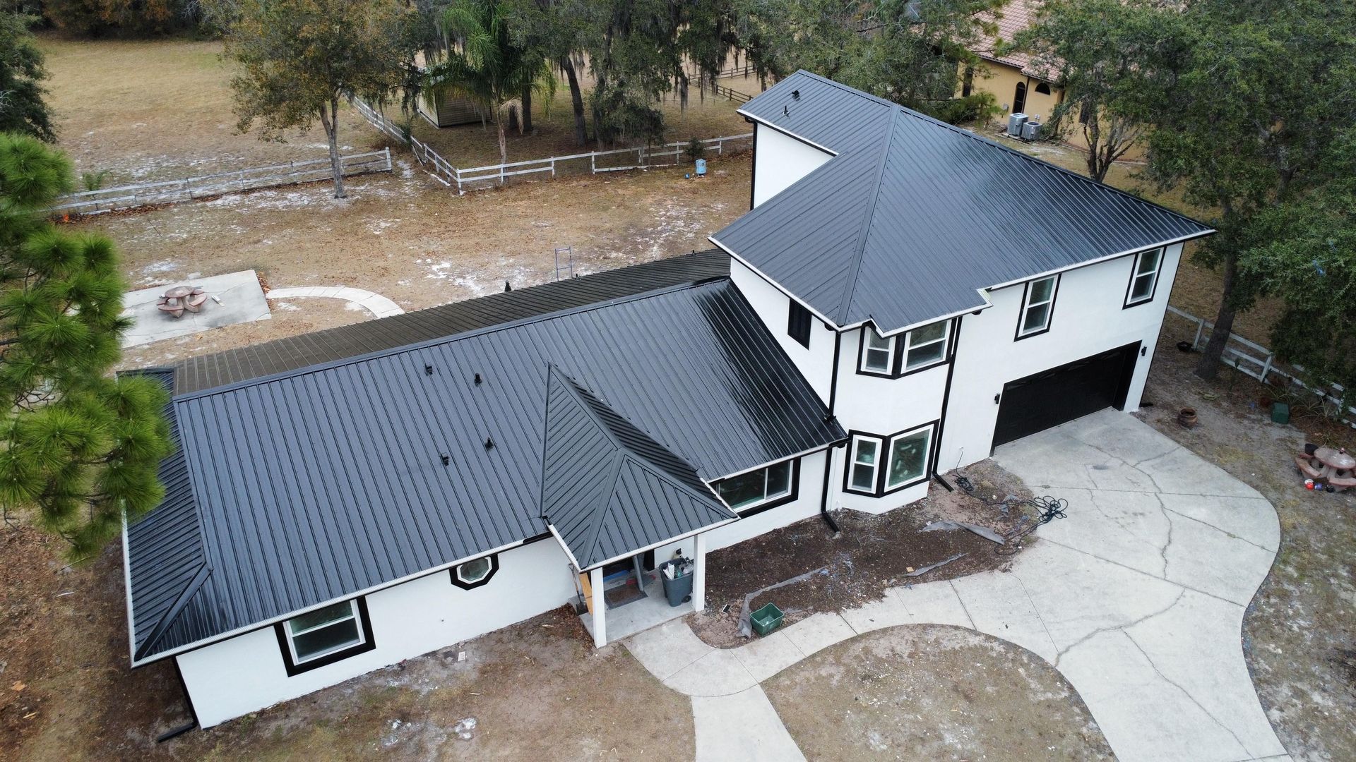 Aerial view of a white two-story house with a dark roof and curved driveway in a wooded yard.