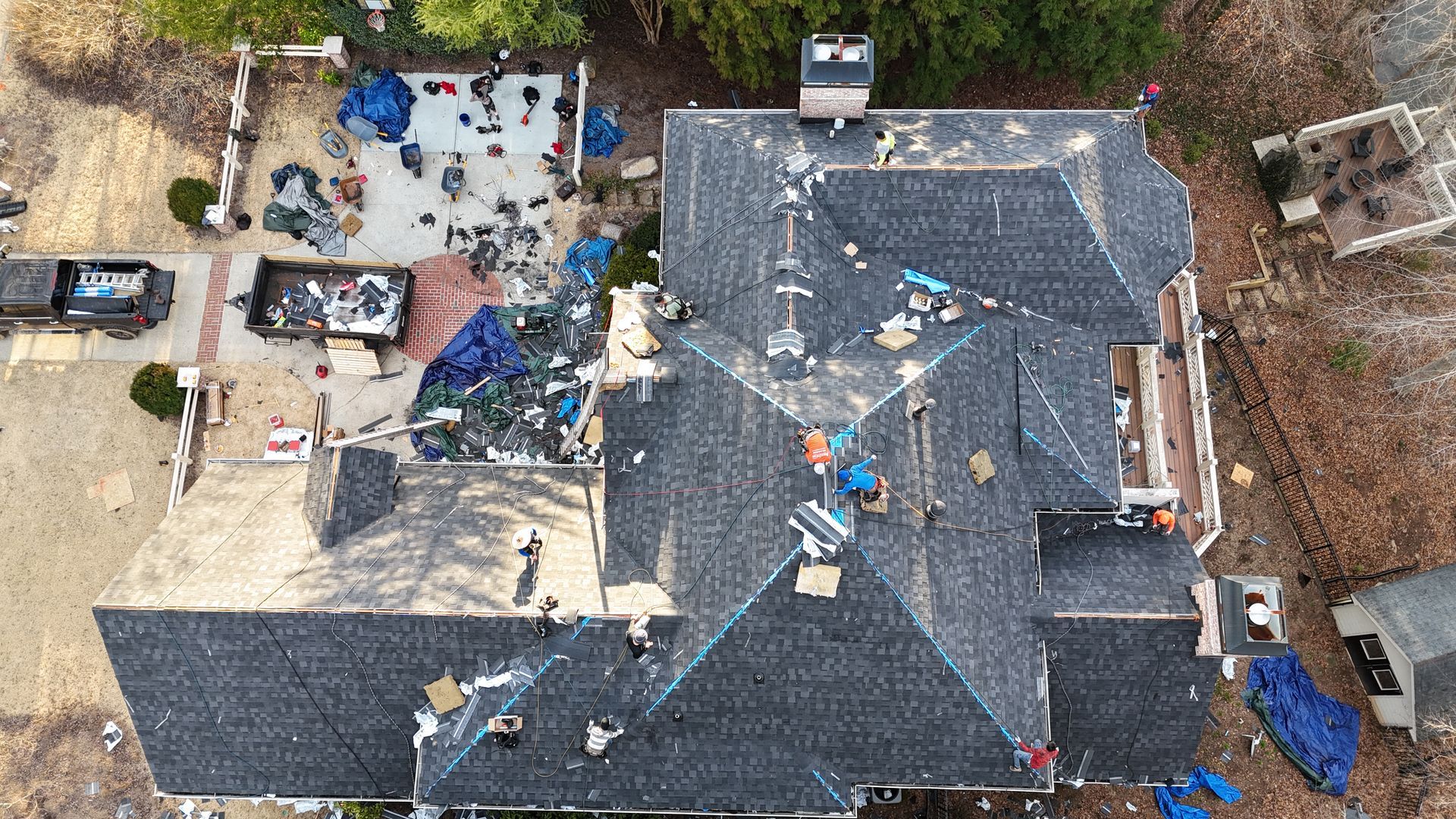 Aerial view of a roof under repair, with workers, blue tarps, and debris around the house.