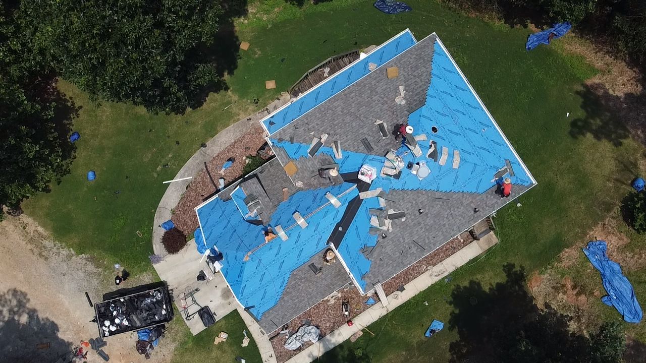 Aerial view of a roof under repair, covered with blue tarps and debris, surrounded by trees.