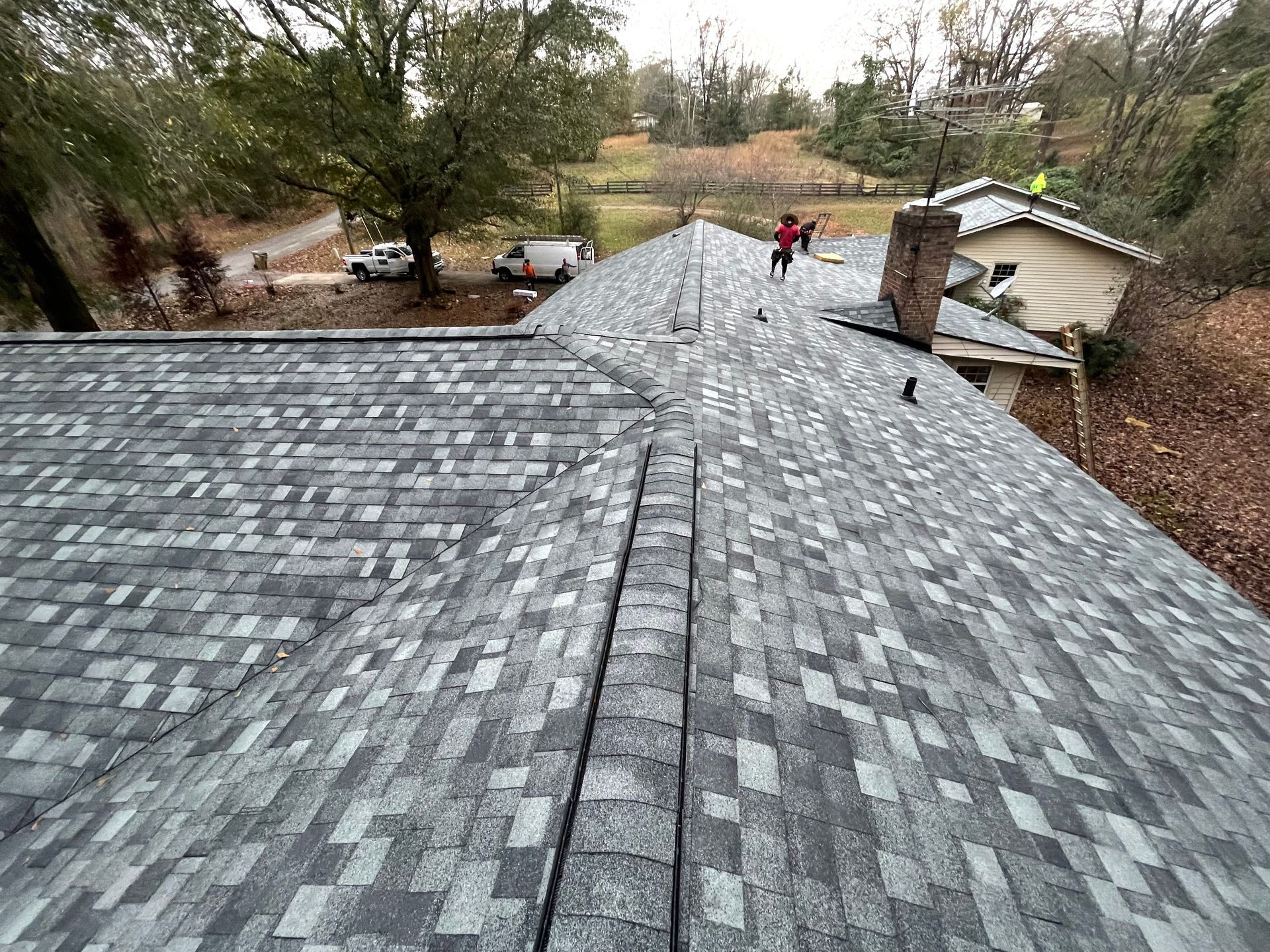 Roof with gray shingles under repair, workers near a shed in a wooded backyard