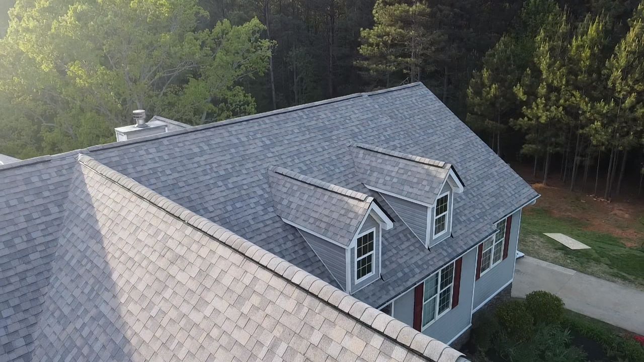 Aerial view of a gray shingled house with dormer windows beside a wooded area