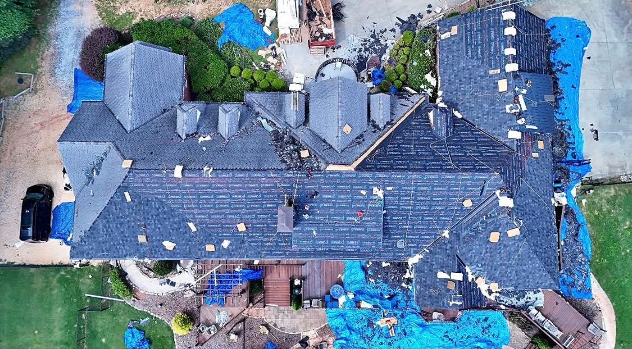 Aerial view of a house with a dark shingled roof, surrounded by debris and blue tarps after storm damage