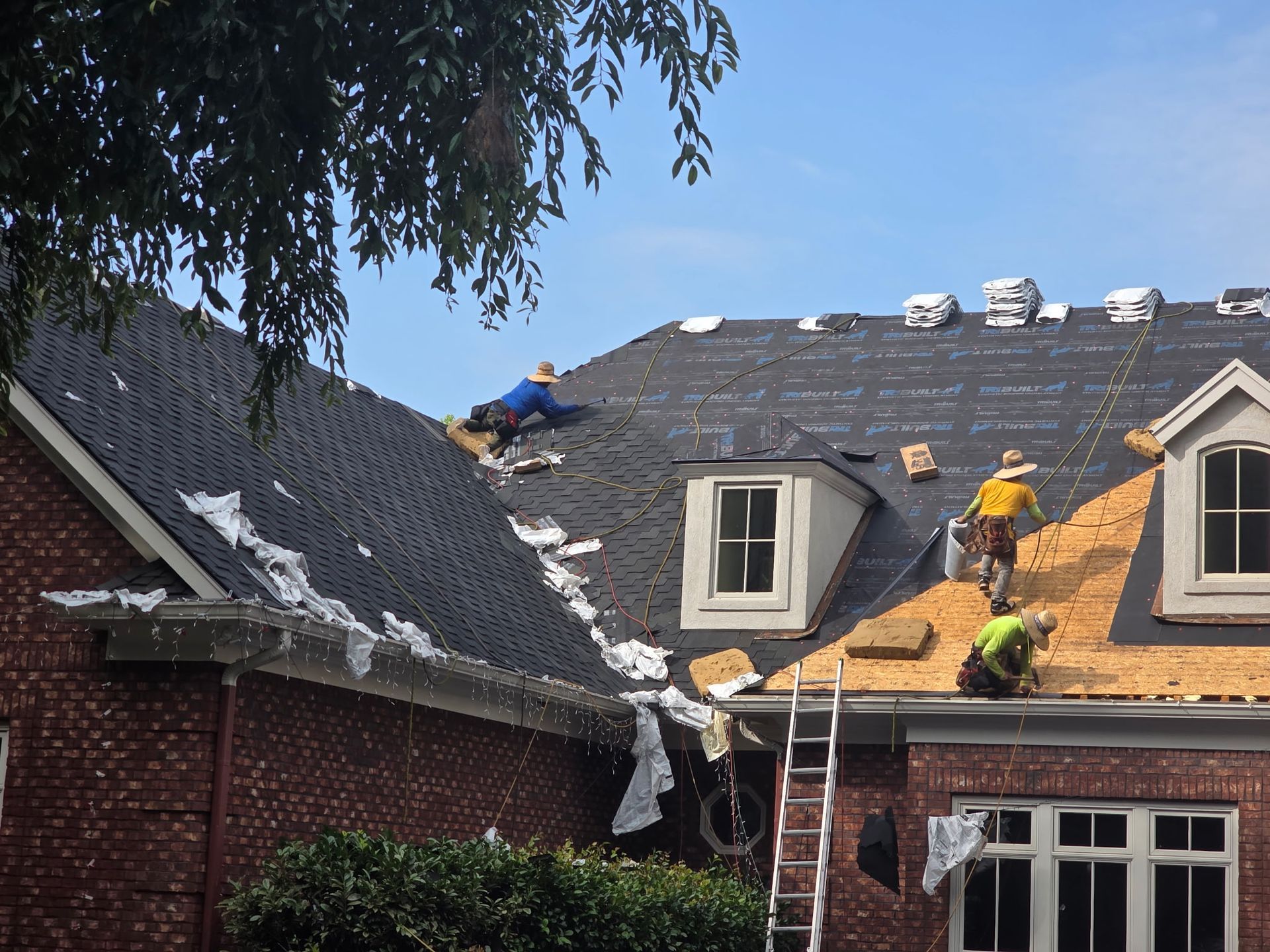 Workers repairing a roof on a brick house, with ladders and debris on the shingles.