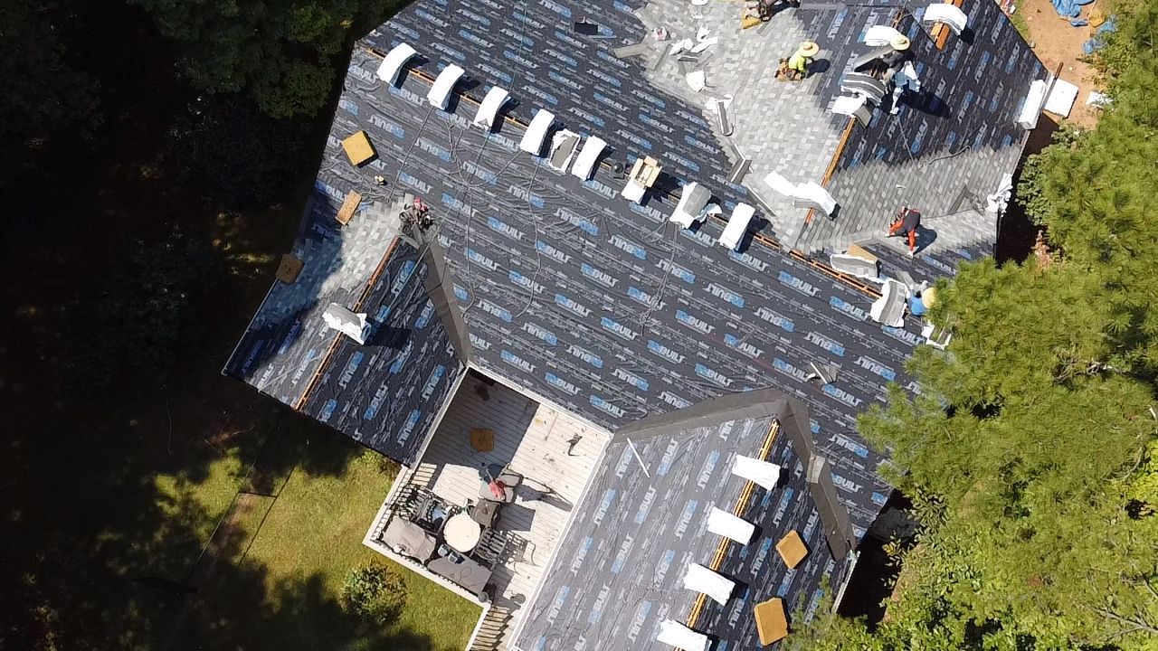 Aerial view of a dark-shingled roof with multiple dormers and a small rooftop terrace surrounded by trees