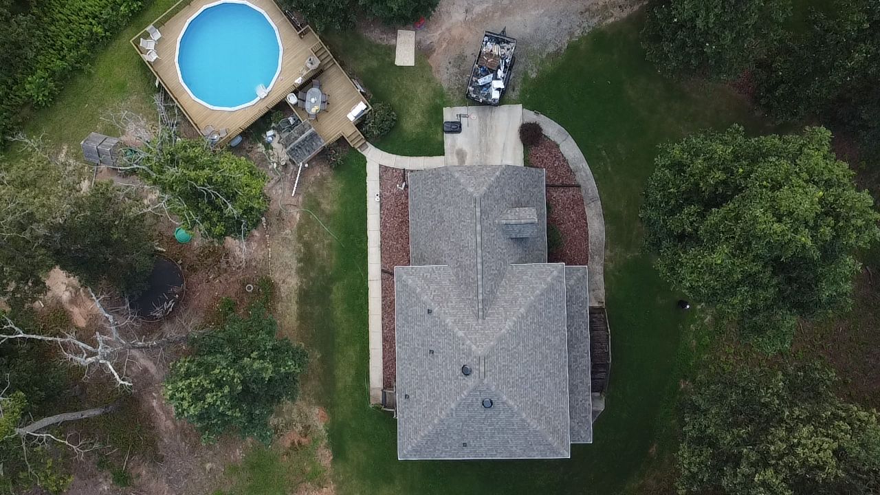 Aerial view of a house with a gray roof, driveway, and a round blue above-ground pool surrounded by trees.