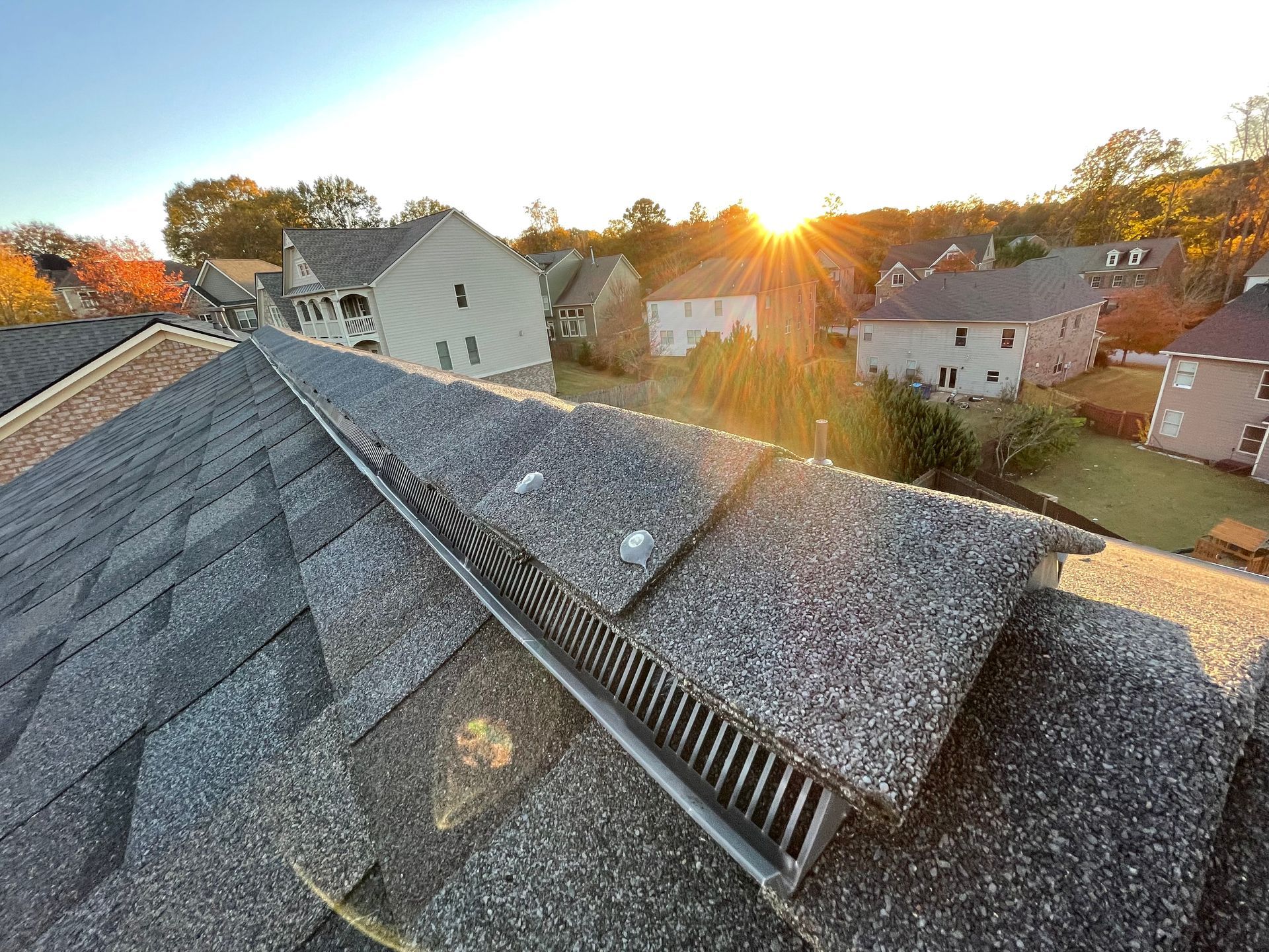 Rooftop at sunset with gray shingles, vent ridge, and houses in a suburban neighborhood below